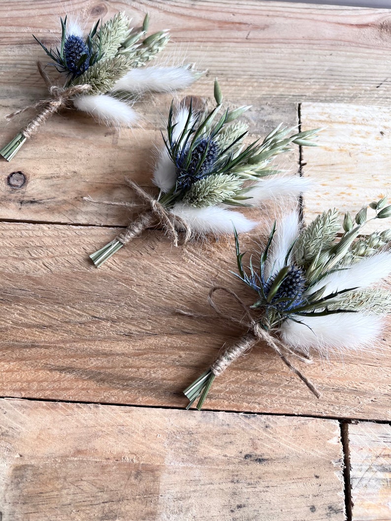 May include: Three small bouquets of dried flowers tied with twine. Each bouquet features blue thistle flowers, white fluffy dried grasses, and green dried grasses.