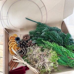 May include: A craft kit for making a wreath, presented in a cardboard box. The kit includes a copper-colored metal wreath frame, fresh greenery, dried orange slices, pine cones, cinnamon sticks, moss, and a burgundy velvet ribbon.