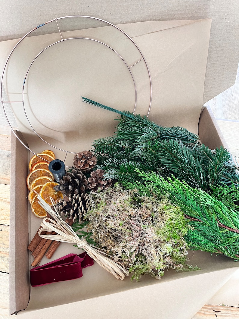 May include: A craft kit for making a wreath, presented in a cardboard box. The kit includes a copper-colored metal wreath frame, fresh greenery, dried orange slices, pine cones, cinnamon sticks, moss, and a burgundy velvet ribbon.