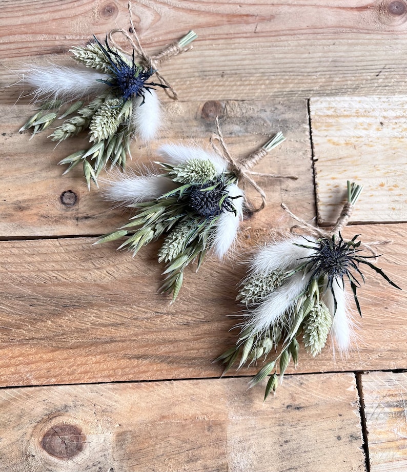 May include: Three small bouquets of dried flowers tied with twine. Each bouquet features dried grasses, white fluffy seed pods, and a dark blue thistle.
