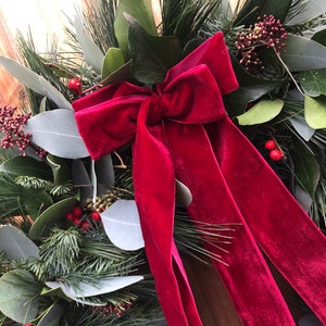 May include: A decorative wreath featuring a deep red velvet bow as the central focal point. The wreath is constructed from a mix of dark green pine needles, broad green leaves, and clusters of small red berries, creating a dense, layered texture.