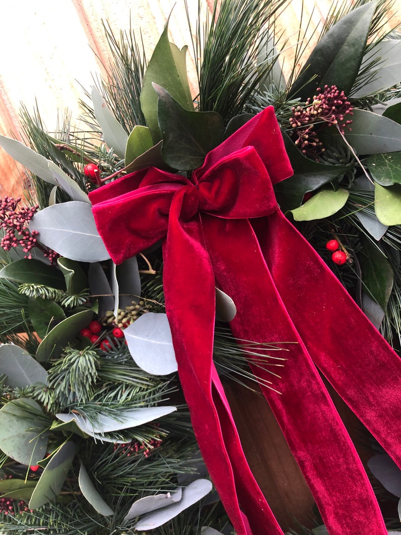 May include: A decorative wreath featuring a deep red velvet bow as the central focal point. The wreath is constructed from a mix of dark green pine needles, broad green leaves, and clusters of small red berries, creating a dense, layered texture.