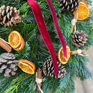May include: A festive Christmas wreath featuring evergreen branches, pine cones, dried orange slices, and cinnamon sticks. A deep red velvet ribbon is tied at the top. The wreath is a natural, rustic decoration.