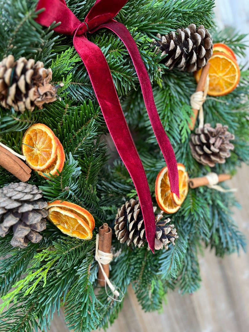 May include: A festive Christmas wreath featuring evergreen branches, pine cones, dried orange slices, and cinnamon sticks. A deep red velvet ribbon is tied at the top. The wreath is a natural, rustic decoration.