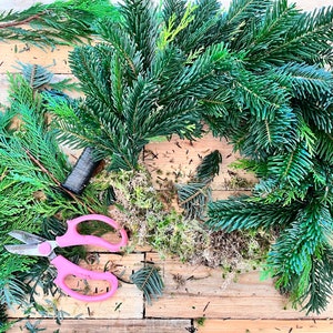 May include: A close-up shot of a Christmas wreath in progress, featuring fresh evergreen branches, moss, and pink handled scissors. The wreath is partially assembled on a wooden surface, with scattered clippings and a spool of wire.