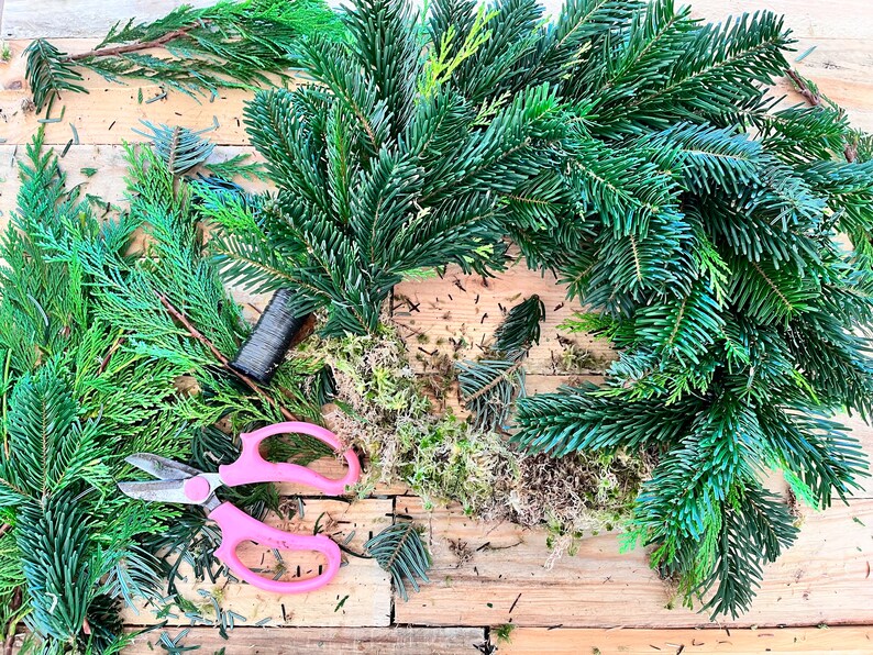 May include: A close-up shot of a Christmas wreath in progress, featuring fresh evergreen branches, moss, and pink handled scissors. The wreath is partially assembled on a wooden surface, with scattered clippings and a spool of wire.