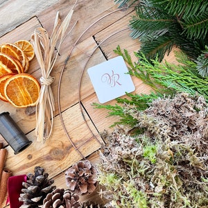 May include: A collection of wreath-making supplies, including a copper wire frame, dried orange slices, cinnamon sticks, pine cones, and greenery. A spool of dark thread and a tag with the initials "VR" are also present. The items are arranged on a wooden surface.