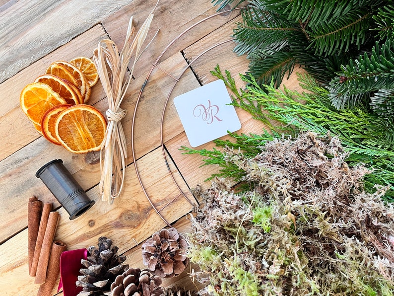 May include: A collection of wreath-making supplies, including a copper wire frame, dried orange slices, cinnamon sticks, pine cones, and greenery. A spool of dark thread and a tag with the initials "VR" are also present. The items are arranged on a wooden surface.