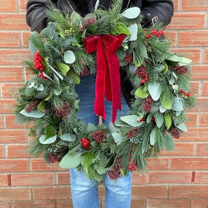 May include: A circular holiday wreath constructed from green pine needles, eucalyptus leaves, and clusters of small red berries. A large, deep red velvet ribbon with long, flowing tails is tied at the top center of the wreath, creating a festive display.