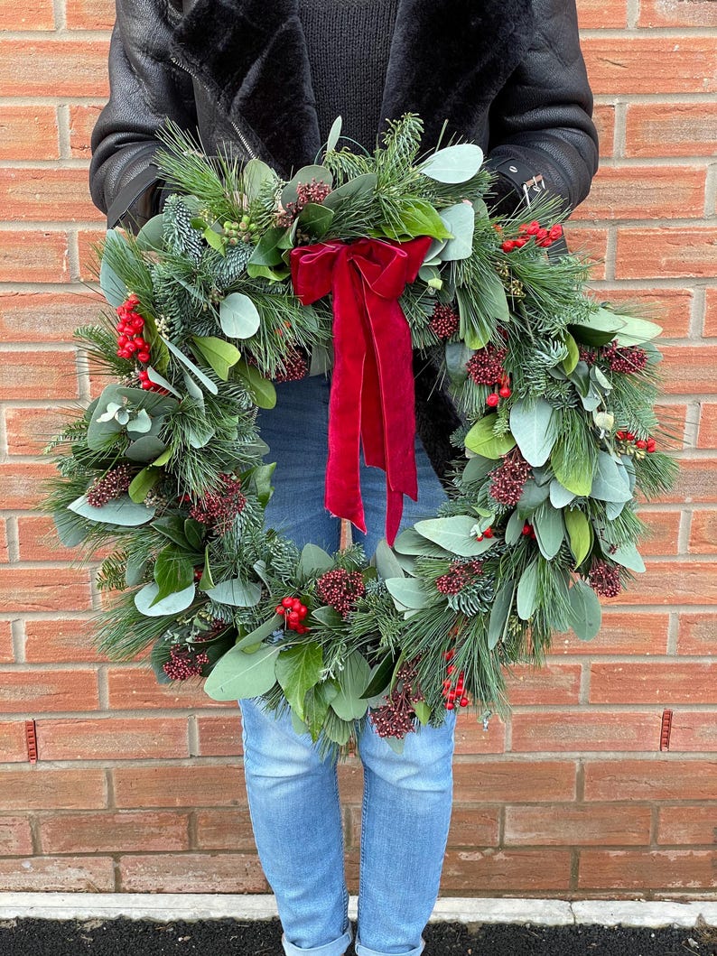 May include: A circular holiday wreath constructed from green pine needles, eucalyptus leaves, and clusters of small red berries. A large, deep red velvet ribbon with long, flowing tails is tied at the top center of the wreath, creating a festive display.