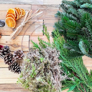 May include: Overhead shot of wreath-making supplies on a wooden surface. Includes a metal ring, fresh evergreen branches, dried orange slices, pine cones, cinnamon sticks, a red velvet ribbon, and floral wire.