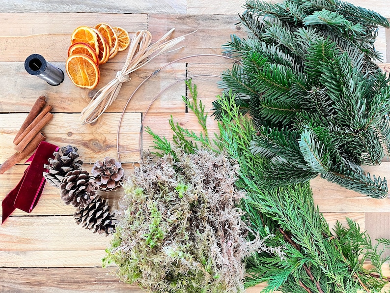 May include: Overhead shot of wreath-making supplies on a wooden surface. Includes a metal ring, fresh evergreen branches, dried orange slices, pine cones, cinnamon sticks, a red velvet ribbon, and floral wire.