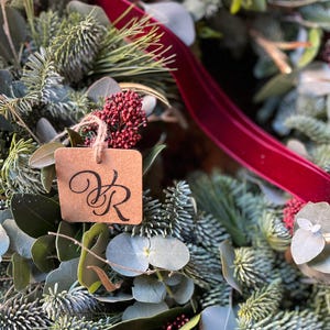 May include: A decorative wreath composed of green pine needles, eucalyptus leaves, and clusters of small red berries. A deep red velvet ribbon is woven through the foliage, and a square brown paper tag with the black printed letters VR hangs.
