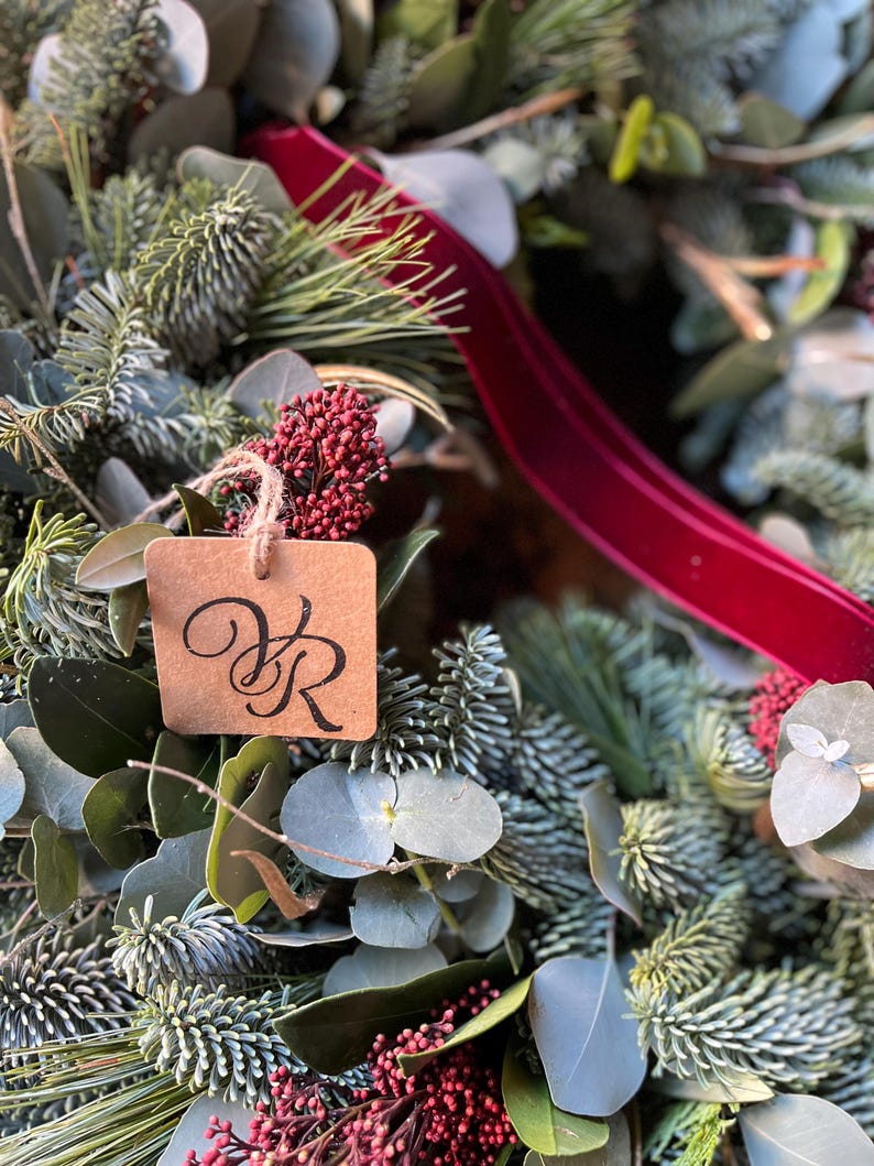 May include: A decorative wreath composed of green pine needles, eucalyptus leaves, and clusters of small red berries. A deep red velvet ribbon is woven through the foliage, and a square brown paper tag with the black printed letters VR hangs.