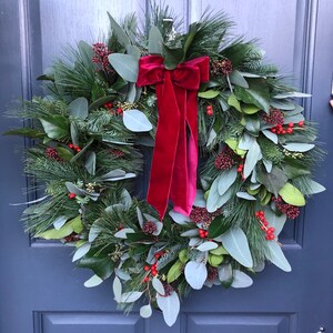 May include: A circular wreath composed of green pine needles, eucalyptus leaves, and magnolia leaves, accented with clusters of small red berries. A large, deep red velvet ribbon with long, flowing tails is tied at the top center of the wreath.