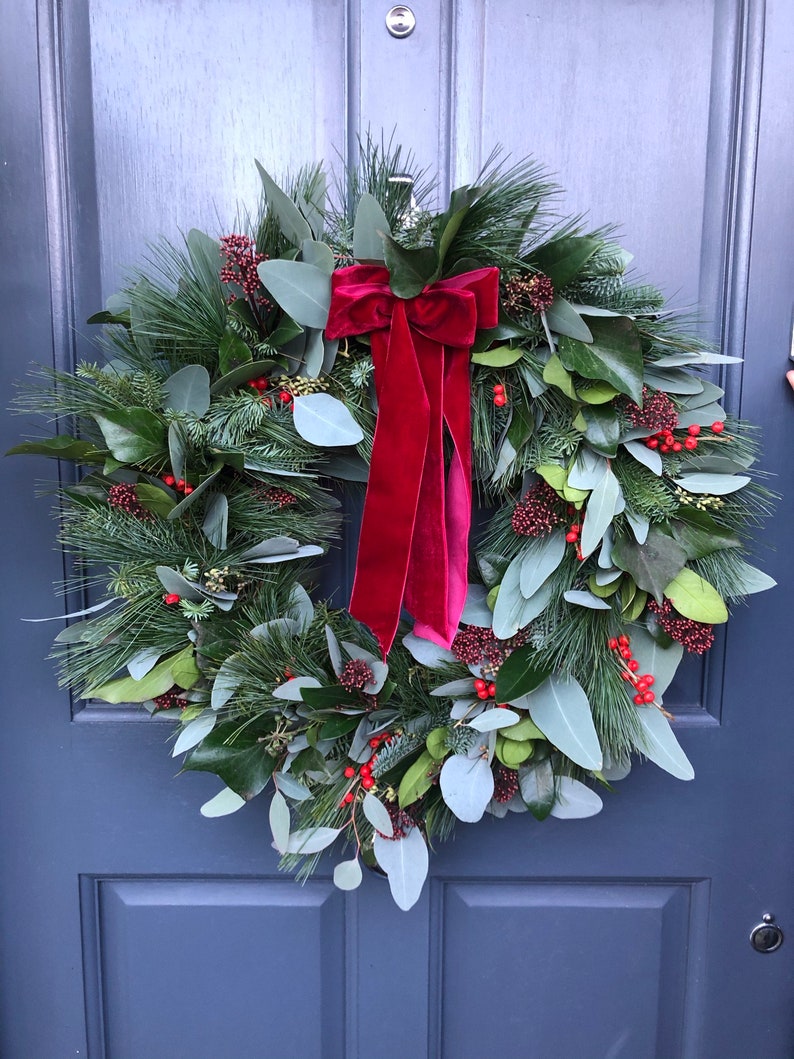 May include: A circular wreath composed of green pine needles, eucalyptus leaves, and magnolia leaves, accented with clusters of small red berries. A large, deep red velvet ribbon with long, flowing tails is tied at the top center of the wreath.