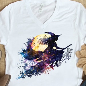 May include: White t-shirt with a black silhouette of a witch flying on a broomstick in front of a full moon. The moon is yellow with a purple and blue galaxy background.