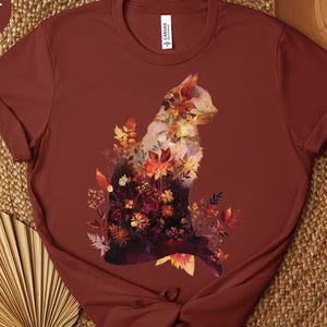 May include: Rust-colored t-shirt featuring a cat silhouette filled with autumn leaves in shades of red, orange, and yellow. The shirt has short sleeves and a crew neck. The word "RUST" is in a circle in the upper left corner.