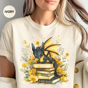 Puede incluir: Camiseta de color marfil con una ilustraci&oacute;n en acuarela de un drag&oacute;n negro con alas amarillas posado sobre una pila de libros. El drag&oacute;n est&aacute; rodeado de flores amarillas y vegetaci&oacute;n. La camiseta tiene un corte holgado.