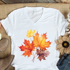 May include: White v-neck t-shirt with a watercolor design of fall leaves in shades of orange, yellow, and brown.