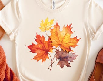 May include: Cream-colored t-shirt featuring a watercolor design of autumn maple leaves in shades of red, orange, and yellow. The shirt is styled with brown sandals and a rust-colored scarf. The word "NATURAL" is printed on a beige circle.