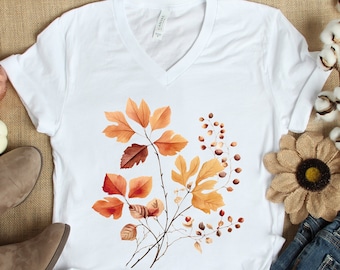 May include: White V-neck t-shirt with a graphic print of orange and brown autumn leaves. The shirt is laid flat on a burlap surface with a pair of brown ankle boots, a small white pumpkin, and a cotton branch with white cotton balls.
