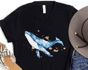 May include: Black V-neck t-shirt featuring a watercolor blue whale design with gold accents. The image also includes a gray hat, denim shorts, and striped sandals, creating a casual, summery aesthetic.