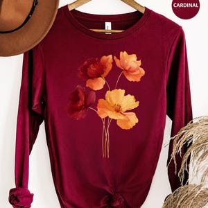 May include: A cardinal red long-sleeve shirt featuring a graphic print of a bouquet of flowers in shades of deep red, orange, and yellow. The shirt has a crew neckline and long sleeves with ribbed cuffs. A brown hat is positioned above the shirt.