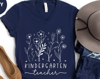 Può includere: Una maglietta blu navy a collo a V con un motivo floreale bianco e il testo "Kindergarten teacher".