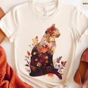 May include: Cream-colored t-shirt featuring a cat silhouette filled with autumn-themed floral patterns in shades of red, orange, and brown. The shirt is styled with a brown hat, orange scarf, and brown sandals. The word "NATURAL" is printed on the top right.