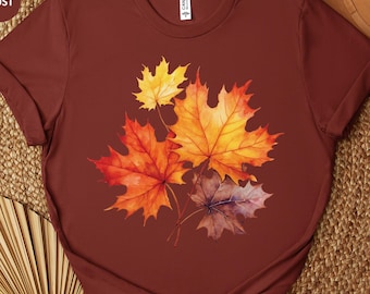 May include: Rust-colored t-shirt featuring a watercolor design of autumn maple leaves in shades of yellow, orange, and red. The shirt has short sleeves and a crew neck. The word "RUST" is printed on the top left.