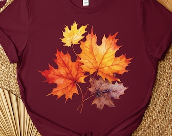 May include: Maroon t-shirt featuring a watercolor design of autumn maple leaves in shades of yellow, orange, and brown. The leaves are clustered together, creating a seasonal fall theme. The shirt is made of a soft material.