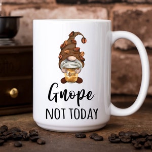 May include: White ceramic coffee mug with a cartoon gnome holding a coffee cup. The gnome is wearing a brown hat with coffee beans. The mug has the text "Gnope Not Today" printed on it.