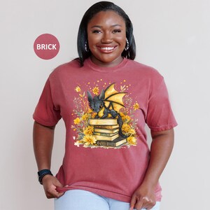 Puede incluir: Camiseta color ladrillo con una ilustraci&oacute;n de un drag&oacute;n negro con alas amarillas sentado sobre libros, rodeado de flores amarillas. La camiseta de manga corta tiene un corte holgado, ideal para el uso diario.