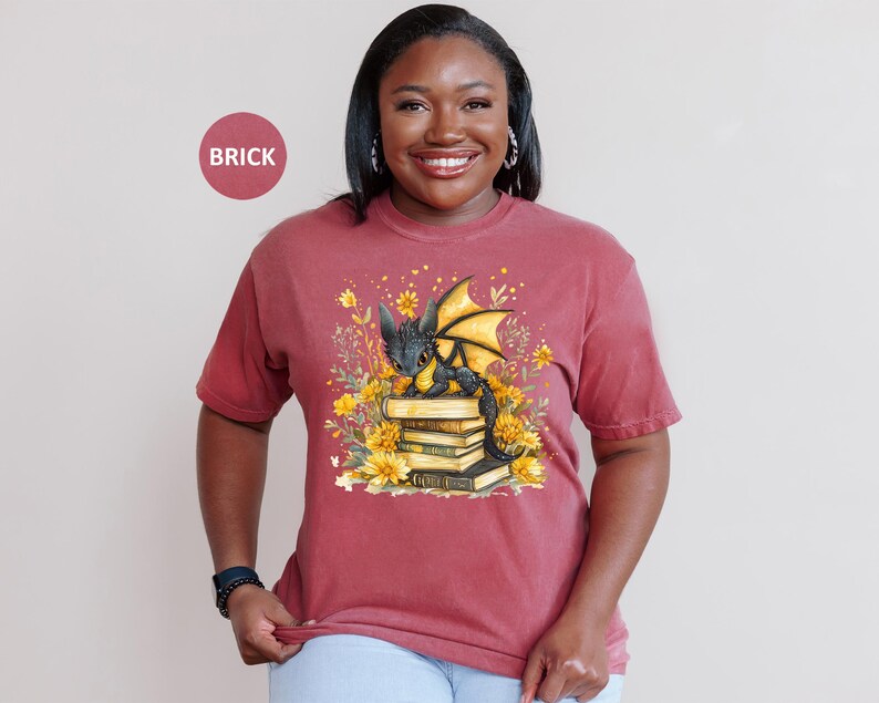 Puede incluir: Camiseta color ladrillo con una ilustraci&oacute;n de un drag&oacute;n negro con alas amarillas sentado sobre libros, rodeado de flores amarillas. La camiseta de manga corta tiene un corte holgado, ideal para el uso diario.