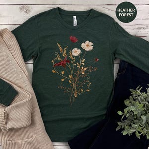 Op de afbeelding: Een donkergroen shirt met lange mouwen met een bloemenontwerp met rode en witte bloemen en goudkleurige stelen. Het shirt wordt gecombineerd met een beige vest en een donkerblauwe jeans. De tekst "Heather Forest" staat in een groene cirkel.