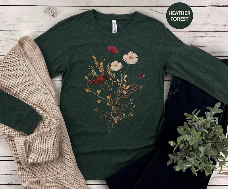Op de afbeelding: Een donkergroen shirt met lange mouwen met een bloemenontwerp met rode en witte bloemen en goudkleurige stelen. Het shirt wordt gecombineerd met een beige vest en een donkerblauwe jeans. De tekst "Heather Forest" staat in een groene cirkel.