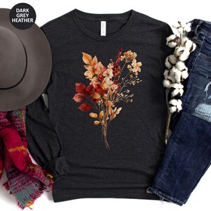 Pu&ograve; includere: Una maglia a maniche lunghe grigio scuro m&eacute;lange con una stampa centrale raffigurante un bouquet di fiori secchi dai toni autunnali, con foglie e fiori color arancione, rosso e beige. &Egrave; presentata insieme a un cappello grigio, una sciarpa a quadri rossa e jeans blu.