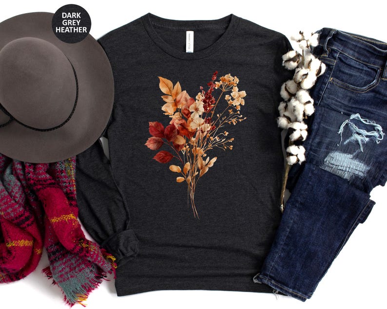 Pu&ograve; includere: Una maglia a maniche lunghe grigio scuro m&eacute;lange con una stampa centrale raffigurante un bouquet di fiori secchi dai toni autunnali, con foglie e fiori color arancione, rosso e beige. &Egrave; presentata insieme a un cappello grigio, una sciarpa a quadri rossa e jeans blu.
