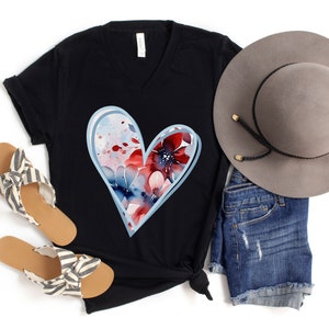 May include: A black t-shirt with a blue and red watercolor heart design. The heart is outlined in light blue and features a floral pattern. The shirt is paired with denim shorts, a gray hat, and striped sandals.