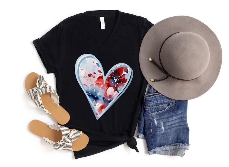 May include: A black t-shirt with a blue and red watercolor heart design. The heart is outlined in light blue and features a floral pattern. The shirt is paired with denim shorts, a gray hat, and striped sandals.