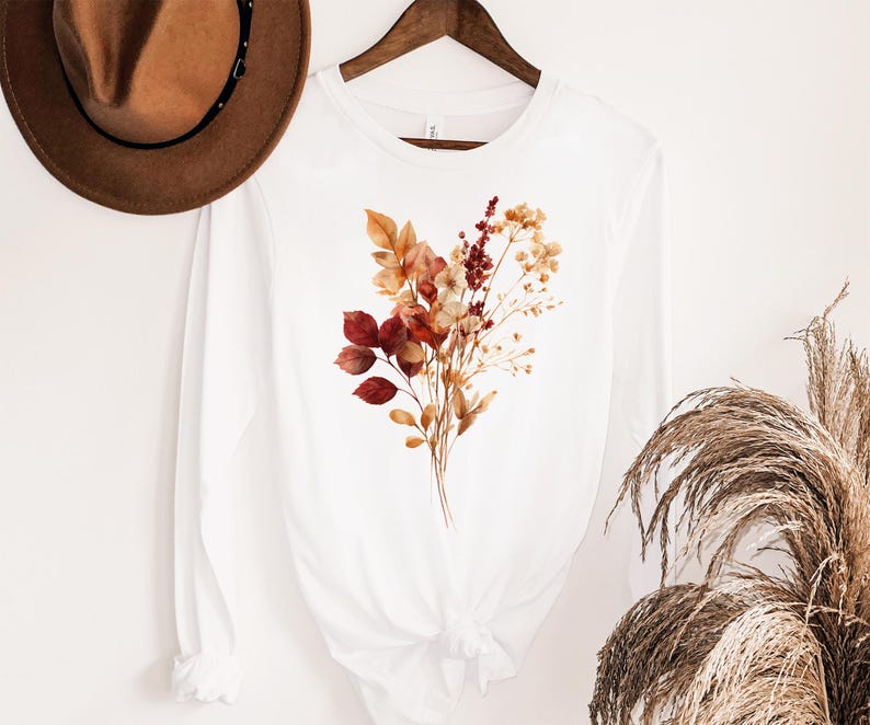 Pu&ograve; includere: Una maglietta bianca a maniche lunghe con una stampa grafica centrale di un bouquet di fiori secchi nei toni del ruggine, arancione e crema. La maglietta &egrave; appesa a una gruccia di legno, con un cappello di feltro marrone sopra la spalla.