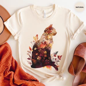May include: Cream-colored t-shirt featuring a cat silhouette filled with autumn-themed floral patterns in shades of red, orange, and brown. The shirt is styled with a brown hat, orange scarf, and brown sandals. The word "NATURAL" is printed on the top right.