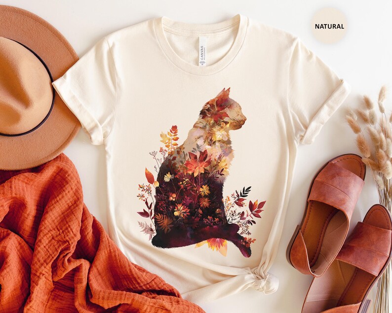 May include: Cream-colored t-shirt featuring a cat silhouette filled with autumn-themed floral patterns in shades of red, orange, and brown. The shirt is styled with a brown hat, orange scarf, and brown sandals. The word "NATURAL" is printed on the top right.