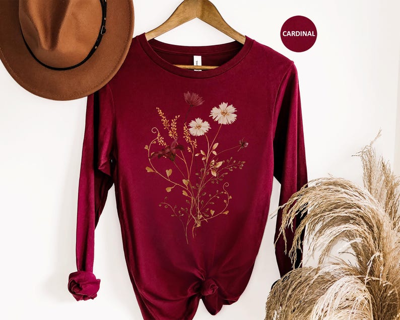 Op de afbeelding: Een lange mouwen, kardinaalrood shirt met een bloemenontwerp. De afbeelding toont witte en bruine bloemen met delicate stelen en bladeren. Het shirt hangt aan een houten hanger. Een bruine hoed is zichtbaar in de linkerbovenhoek. Het woord "CARDINAL" staat in een rode cirkel.