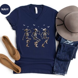 May include: A navy blue v-neck t-shirt with a graphic of three skeletons wearing cowboy hats and dancing. The shirt is paired with a brown wide-brimmed hat and blue jeans.