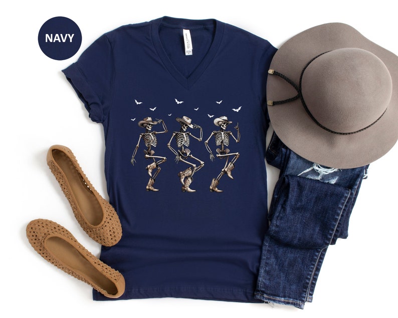 May include: A navy blue v-neck t-shirt with a graphic of three skeletons wearing cowboy hats and dancing. The shirt is paired with a brown wide-brimmed hat and blue jeans.