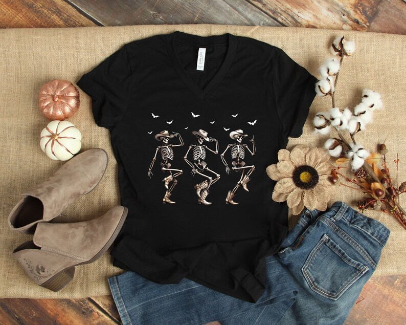 May include: Black v-neck t-shirt with a graphic of three skeletons wearing cowboy hats and dancing. The skeletons are white with black outlines. The background is black with white bats.