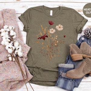 May include: A heather olive t-shirt with a floral print featuring red, white, and gold flowers. The shirt is shown with a pair of jeans and a sweater.