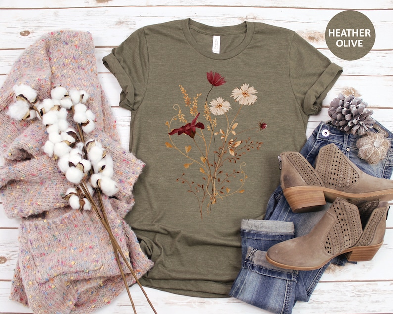 May include: A heather olive t-shirt with a floral print featuring red, white, and gold flowers. The shirt is shown with a pair of jeans and a sweater.